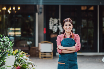 Smiling female cafe owner in apron standing confidently outside her coffee shop with arms crossed,...