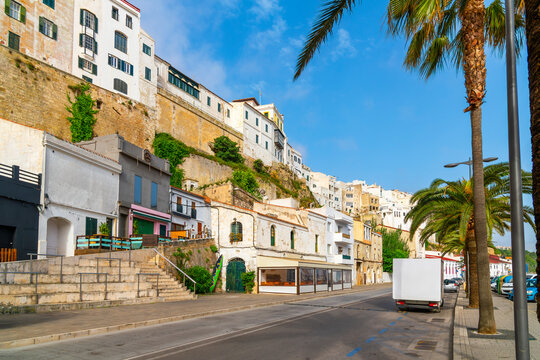 The whitewashed cliffside buildings with sidewalk cafes and shops below along the waterfront at the port harbor of Mahon, on the Mediterranean Balearic island of Menorca, Spain.