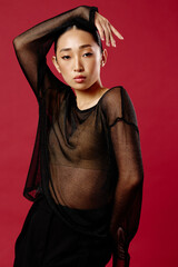 Fashion asian woman in black sheer top and pants posing in front of a red background with hands on...