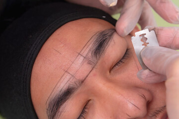 Using a razor to shape the edges of the eyebrow. During a microblading or embroidery procedure for men at an aesthetic clinic.