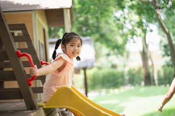 Asian little girl is sitting on a green slide at a playground. The playground is full of colorful...