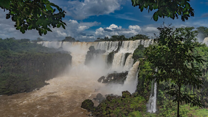 A picturesque cascade of waterfalls. Streams of white foaming water collapse from the rocks into the riverbed. Spray and fog rise into the air. Lush tropical vegetation. Blue sky, clouds. Iguazu Falls