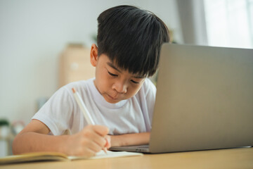 Naklejka premium A young boy is sitting at a table with a laptop and a pencil. He is writing something on a piece of paper. The scene suggests that the boy is engaged in a creative or educational activity