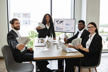 Diverse business team celebrating financial success in office. Colleagues holding money, smiling, and displaying graphs and charts. Professionals in formal attire enjoying successful business results.
