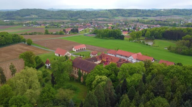 A drone moves forward, Kalsdorf Castle, green fields and trees under cloudy skies.