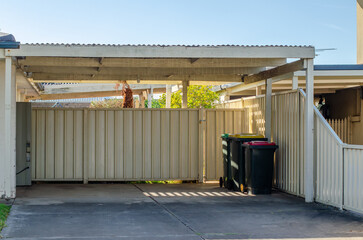 An open and accessible carport is attached to a residential suburban house to provide some shade and shelter, without a gate. Household rubbish bins are stored inside.