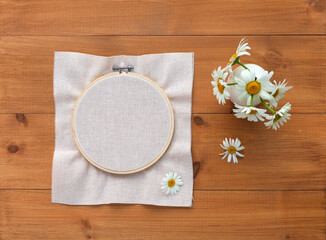 Summer crafts. Light canvas is stretched into hoop for hand embroidery with cross stitch, satin stitch or satin ribbons. Nearby bouquet of daisies on wooden table. Flat lay, copy space, mockup