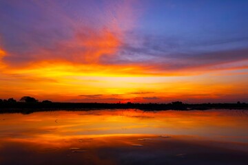 Scenic view of beautiful sunrise or dawn above the pond or lake at spring or early summer morning with cloudy sky background, clouds over water and reed grass with dew at foreground. Water reflection.