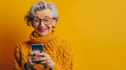 Fototapeta premium Senior woman with gray hair and glasses smiling while using smartphone. Wearing a mustard yellow sweater and standing against a vibrant yellow background