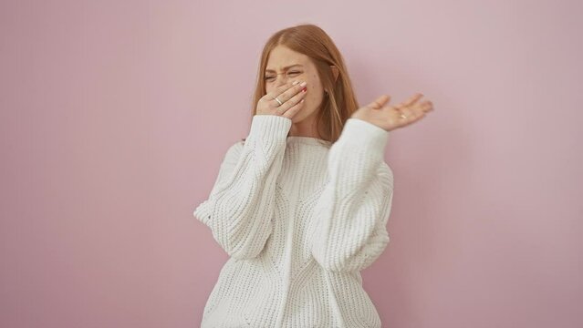 Redhead young woman holding breath in disgust, repulsed by a terrible stink, smelling an intolerable, stinky bad smell over a pink isolated background while standing, wearing a sweater.