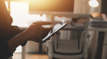 Obraz premium Smiling businesswoman using her phone in the office. Small business entrepreneur looking at her mobile phone and smiling while communicating with her office colleagues