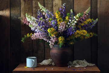 dark rural still life with lupines in a clay jug on the table. garden flowers and rustic interior.