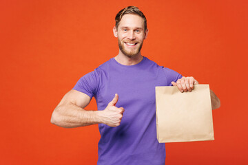 Young man wear purple t-shirt casual clothes hold clear blank craft paper takeaway bag show thumb up isolated on red orange background. Food products delivery courier service from shop or restaurant.