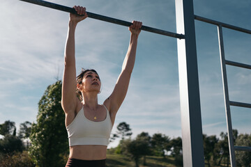 Determined calisthenic woman performing pull-ups at outdoor fitness park. Young athletic woman engaging in a pull-up workout on horizontal bars under a clear sky
