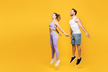 Full body side profile view young strong fitness trainer sporty two man woman wearing blue clothes spend time in home gym jumping high isolated on plain yellow background. Workout sport fit concept.