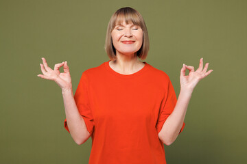 Elderly blonde woman 50s years old wear orange t-shirt casual clothes spreading hands in yoga om aum gesture relax meditate try calm down isolated on plain pastel green background. Lifestyle concept.