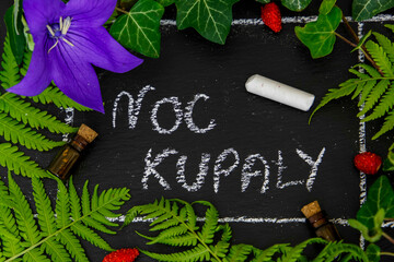 Napis kredom Noc Kupały na czarnym tle w otoczeniu liści paproci, bluszczu, poziomek i kwiatu. 