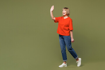 Full body side view smiling elderly blonde woman 50s years old she wear orange t-shirt casual clothes waving hand walk go isolated on plain pastel green background studio portrait. Lifestyle concept.