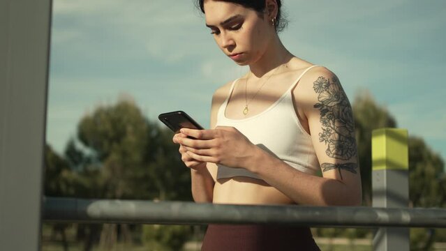Young woman using her phone in a sunny street workout park.