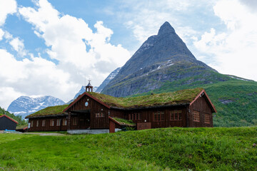 In Norway, a rustic wooden cabin with a grassy roof is nestled among lush green mountains, creating...
