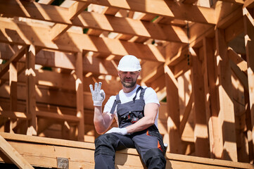 Man manager on construction site of wooden-framed house gestures the OK sign. The concept pertains to modern ecological construction.
