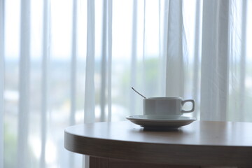 A white mug sits atop a wooden table next to a bright window, invitingly filled with tea drink. Concept Cozy Indoor Table.