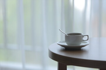 A white mug sits atop a wooden table next to a bright window, invitingly filled with tea drink. Concept Cozy Indoor Table.