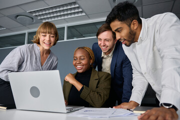 Four happy young adults in businesswear working on laptop at desk in office