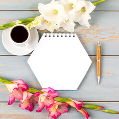 Composition with opened blank notepad and pen, cup of black coffee, gladiolus flowers on blue wooden table. Festive office desktop concept. Morning coffee cup.