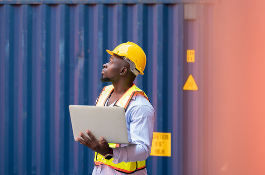 Male African engineer worker wearing yellow hard hat and reflective safety vest using laptop computer for container inspection at cargo shipyard. Transportation import and export logistic industry con - Powered by Adobe