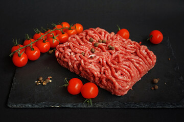 Fresh minced meat on a black slate tray next to cherry tomatoes and spices on a dark background