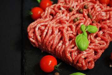 Fresh minced meat on a black slate tray next to cherry tomatoes, basil and spices on a dark background