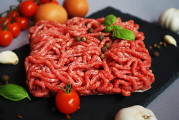 Fresh minced meat on a black slate tray next to cherry tomatoes, basil, eggs and spices on a gray background