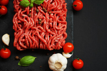 Fresh minced meat on a black slate tray next to garlic, cherry tomatoes, basil and spices on a dark...