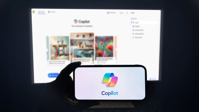 Copilot app running on a modern mobile phone. The smartphone is being held in landscape mode, in the darkness. A PC monitor with the Copilot website in the background.