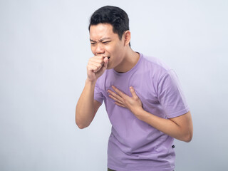 Young Asian man in lavender t-shirt coughing into his fist while holding his chest, indicating discomfort or illness. Studio shot on plain background capturing his casual outfit