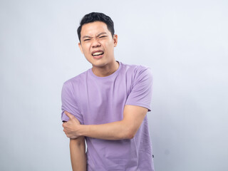 Fototapeta premium Young Asian man in a light purple shirt, wincing in pain while holding his left arm, standing against a plain light grey background, conveying discomfort or injury.