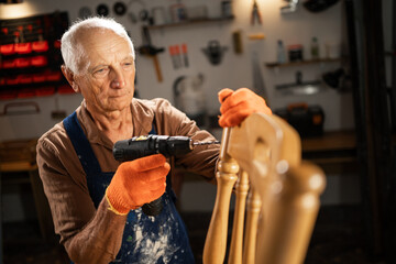 An elderly man drills a hole in furniture, restoration and DIY concept.
