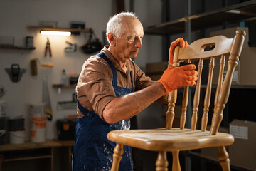 An elderly man in orange gloves and a blue apron sands an old wooden chair in his workshop. He is...
