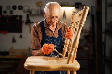 Restoration and repair of an old wooden chair. An elderly man in orange gloves, a blue apron, stands in his workshop and carefully paints furniture with a brush