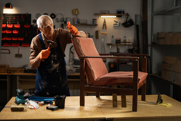 Obraz premium An elderly man in orange gloves and a blue apron begins to restore an old chair in a workshop. He holds a hammer and carefully removes the old fabric. concept second life of old furniture. Copy space.