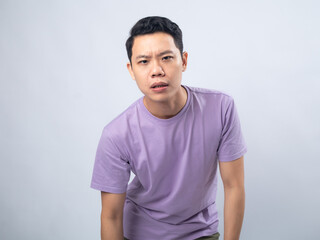Young Asian man in a lavender t-shirt grimacing in pain and holding his lower back. Studio shot on...