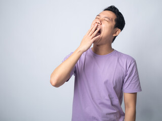 Young Asian man in a lavender t-shirt yawning and covering his mouth with his hand. Studio shot on a plain background, highlighting his sleepy expression and casual fashion.