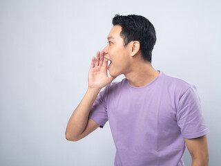 Young Asian man in a lavender t-shirt smiling and shouting with his hand cupped around his mouth. Studio shot on a plain background, highlighting his cheerful expression and casual fashion.