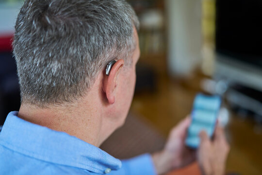 Close Up Of Mature Man Wearing Wireless Hearing Aid Using Mobile Phone  At Home