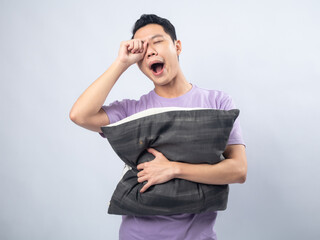 Young Asian man in a lavender t-shirt yawning and rubbing his eye while holding a pillow. Studio shot on a plain background, highlighting his sleepy expression and casual fashion.