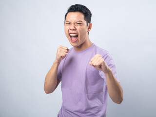 Fototapeta premium Young Asian man in a lavender t-shirt standing with fists raised, shouting energetically. Studio shot on a plain background emphasizing his excited and victorious expression and casual attire