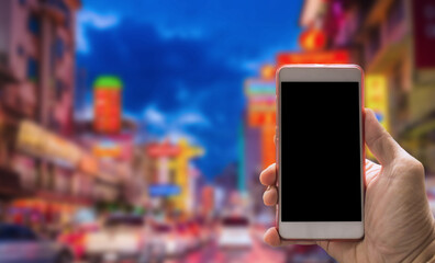 A hand holding a smartphone with a blank screen in front of a vibrant, blurry city street filled...