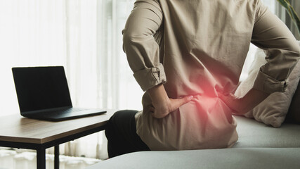 Working businessman sitting on the sofa in the living room at home He used both hands to press down on the lower back. He is suffering from back pain from sitting for a long time.