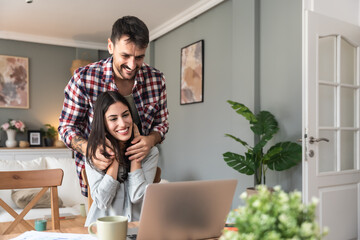 Young introvert freelance business woman trying to work from home office on laptop, nervous for video call communication while her boyfriend trying to cheer her up making faces and fun behind her back